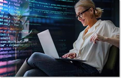 smiling blond woman in glasses working on a laptop with code projected on the wall behind her