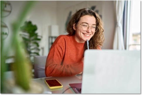 Smiling person in glasses and an orange sweater working at an open laptop